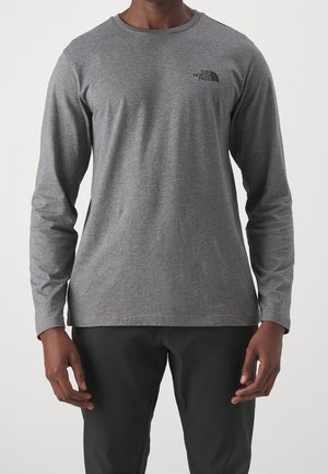 Personne portant un t-shirt gris à manches longues avec col rond et le logo The North Face sur la poitrine, ainsi qu’un pantalon noir, debout devant un fond uni.