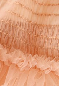 Persikofärgad tulle-tyg med samlad textur och volangkanter. Mjuk och genomskinlig med horisontella ränder och en delikat sömdetalj.