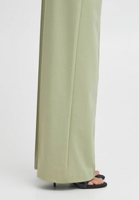 Bledi zeleni široki hlače iz mehkega materiala, z ravnim krojem in razporki, v kombinaciji s črnimi sandali z odprtimi prsti.