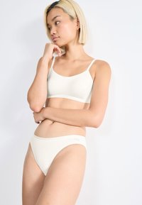 Jonge vrouw met kort blond haar, gekleed in een witte bralette en hoog uitgesneden ondergoed, poseert nadenkend tegen een witte achtergrond.