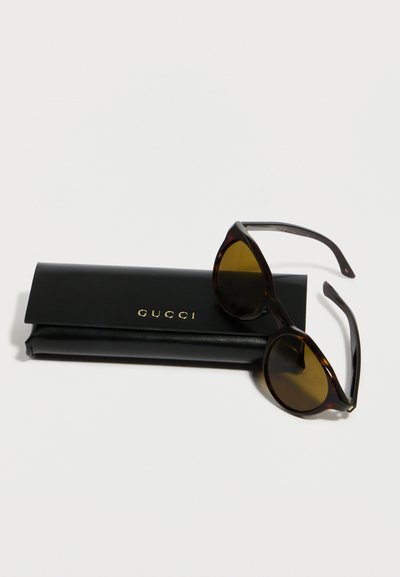 Lunettes de soleil rondes en écaille de tortue avec des verres marron reposant sur un étui Gucci noir avec des lettres dorées sur un fond blanc.