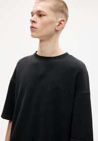 Černá oversized mikina s žebrovaným kulatým výstřihem, krátkými rukávy a jemně strukturovanou tkaninou, s malým vyšívaným logem na hrudi.
