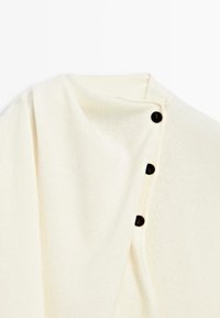 Pull en maille couleur crème avec fermeture latérale à boutons, présentant trois petits boutons foncés le long de la couture de l’épaule.