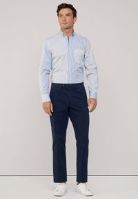 Camisa de botones a rayas en azul claro y blanco, combinada con pantalones chinos azul marino y zapatillas blancas. La camisa tiene un bolsillo en el pecho y mangas largas.