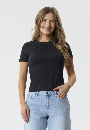 Jeune femme souriante, portant un t-shirt noir à manches courtes et un jean bleu clair à petits pois, debout devant un fond uni.