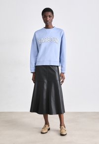 Világoskék pulóver, amelyen a "BARBOUR" felirat szerepel, fekete műbőr midi szoknya és bézs slip-on cipők. Egyszerű dizájn, laza szabású.