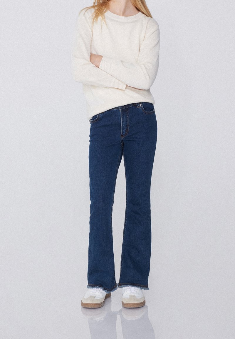 Jeune personne portant un pull blanc, un jean bleu et des baskets blanches, debout les bras croisés devant un fond uni.