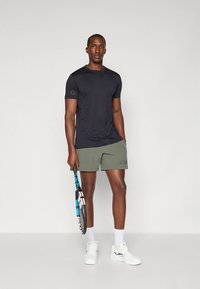 T-shirt desportivo preto, calções verde-oliva com "JORD" impresso, ténis brancos, segurando uma raquete de ténis com um design azul e preto.