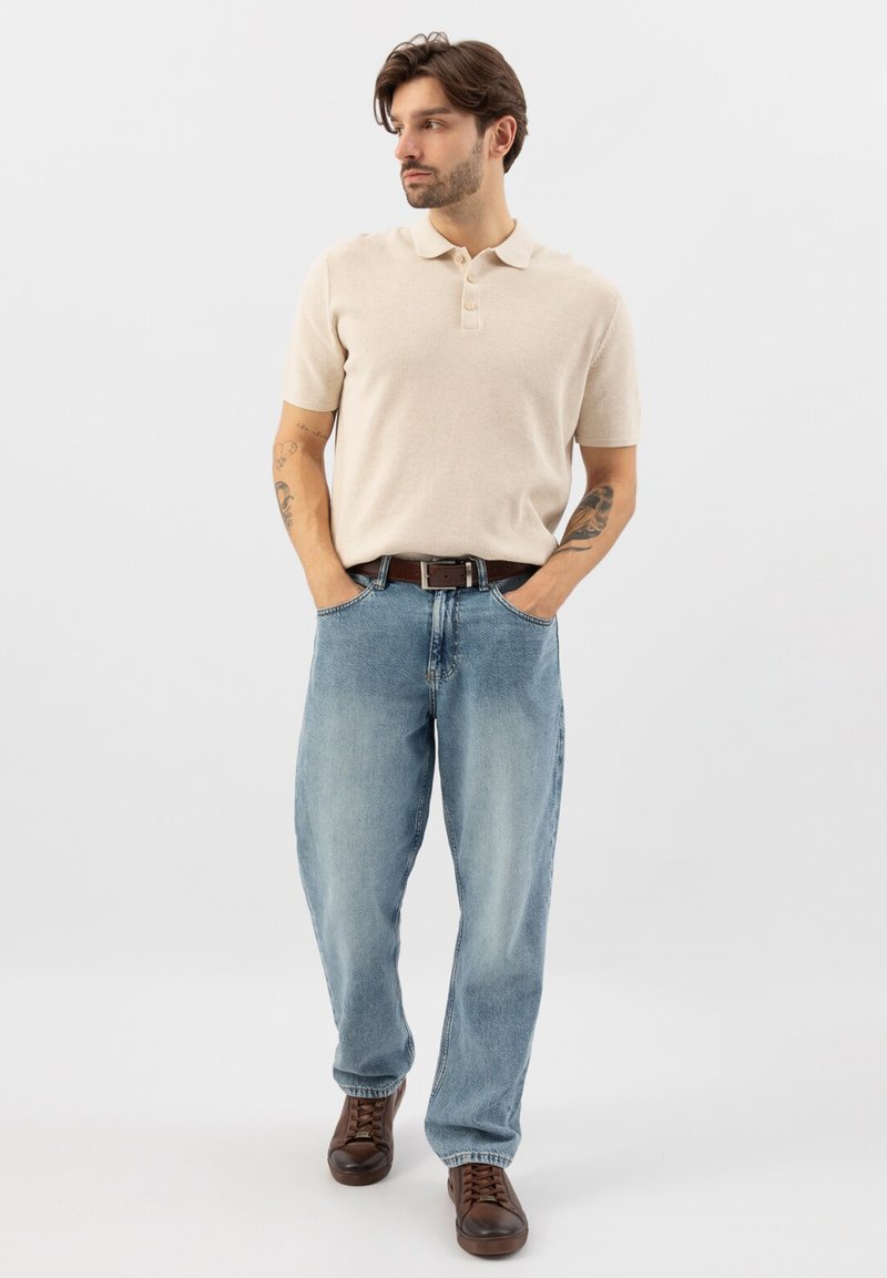 Uomo in piedi con tatuaggi su entrambe le braccia, che indossa una polo beige a maniche corte, jeans azzurri, cintura marrone e scarpe marroni con lacci.