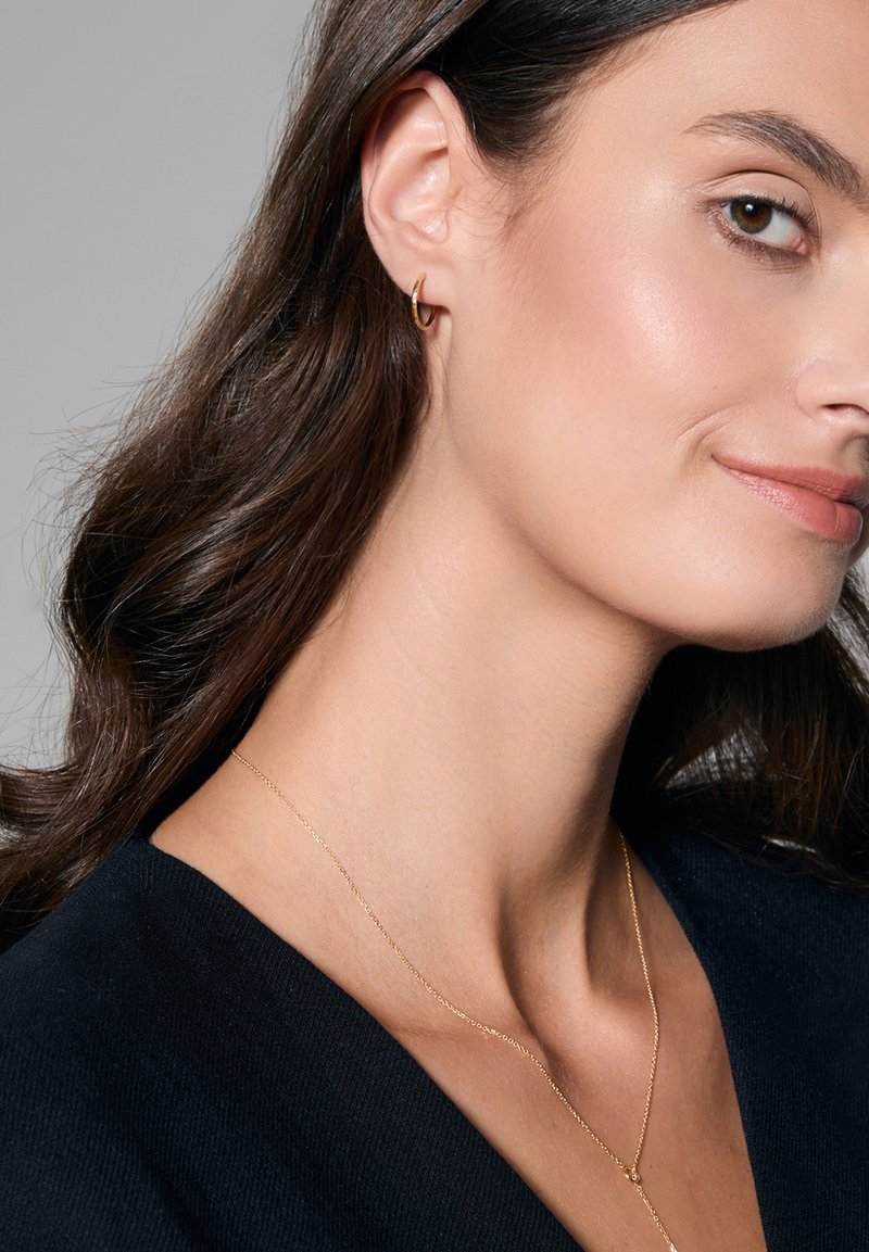 Złoty kolczyk w kształcie koła noszony w lewej uchu, z cienkim złotym naszyjnikiem i ciemnym swetrem. Gładkie włosy opadają na ramiona.