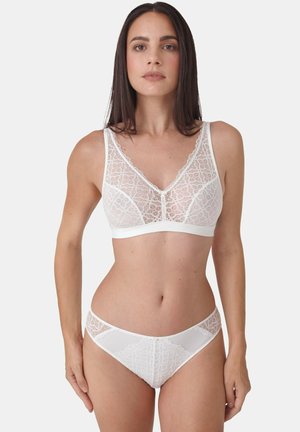 Woman wearing matching white lace bra and panties, standing against plain light background, facing forward with neutral expression.