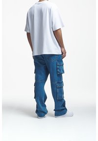 Muž v bílém oversized tričku, volných modrých cargo džínách s několika kapsami a bílých teniskách, stojící na bílém pozadí.