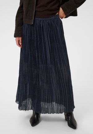 Femme portant une jupe longue plissée foncée, des bottes noires pointues et une veste sombre, une main dans la poche, sur un fond uni.
