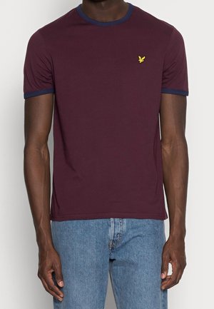 Mand iført en mørkerød t-shirt med korte ærmer med marineblå kant og et lille gult fuglelogo, kombineret med lyseblå jeans.