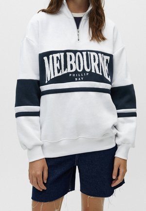 Biała i granatowa bluza z suwakiem do połowy z napisem "MELBOURNE PHILLIP BAY", noszona z ciemnoniebieskimi jeansowymi szortami.