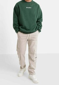 Grüner Pullover mit weißem Text, beige Cargohose mit Seitentaschen und Druckknöpfen, getragen mit weißen Turnschuhen. Weicher Stoff, lässige Passform.