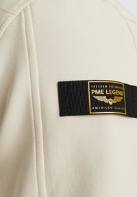 Un tissu beige avec une texture lisse présente une étiquette tissée noire avec un texte doré, indiquant "PME LEGEND AMERICAN CLASSIC" et des graphiques d'ailes.