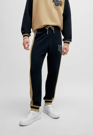 Navy and beige tracksuit with elastic waistband and cuffs. Features logo on chest and left pant leg, complemented by white sneakers.