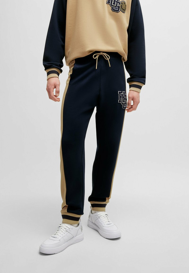 Tuta navy e beige con cintura elastica e polsini. Presenta il logo sul petto e sulla gamba sinistra dei pantaloni, abbinata a sneakers bianche.
