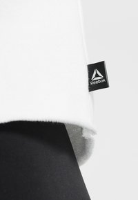 Bílý sportovní tričko s černou nášivkou loga Reebok na lemu, spárované s černými strukturovanými legínami, které zdůrazňují kontrast materiálu a barev.