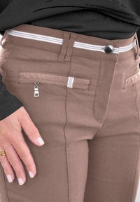 Braune Baumwollmischungshose im Slim-Fit, mit weißen gestreiften Akzenten und einer Reißverschlusstasche. Strukturierter Stoff und sichtbarer Knopfverschluss.