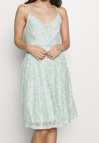 Mintgrön spetsklänning med v-ringning, delikata scallop-kant, figurnära midja och flödande kjol. Har blommig spetsmönster och justerbara axelremmar.