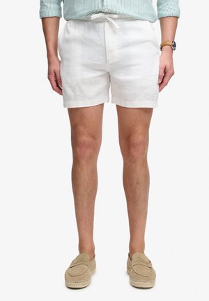 Mężczyzna ubrany w białe szorty ze ściągaczem, zieloną koszulę w paski z podwiniętymi rękawami, beżowe buty wsuwane oraz brązowy zegarek na nadgarstku, pokazany od pasa w dół.