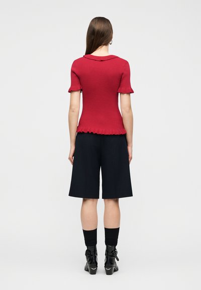 Mujer vistiendo un top de punto rojo de manga corta con bordes con volantes, pantalones cortos negros hasta la rodilla, calcetines negros y botines negros, de pie de espaldas.