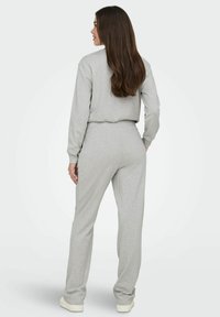 Vrouw met lang bruin haar in een lichtgrijze bijpassende sweatshirt en broek, rechtopstaande met de rug naar de camera tegen een effen achtergrond.