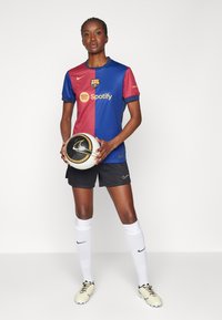 FC Barcelonan pelipaita sinisenä ja maroonina, jossa "Spotify"-logo, yhdistettynä mustiin shortseihin, valkoisiin sukkiin ja pidellen mustaa ja kultaa jalkapalloa.