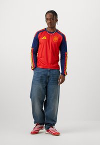 Man som står och bär en röd och blå långärmad Spanska fotbollströjan, lösa blå jeans, röda och vita sneakers samt ett silverkedjehalsband.