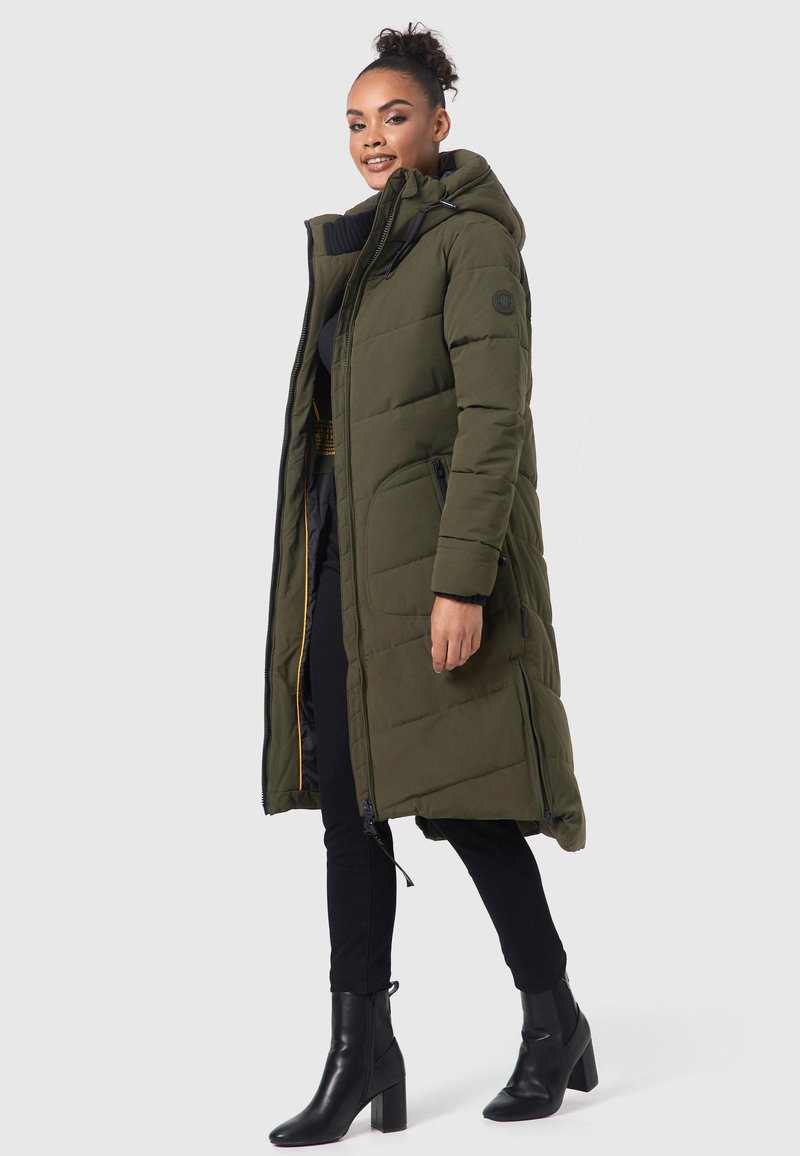Long manteau olive vert matelassé avec capuche, doté d'une fermeture éclair, de poches latérales et d'une doublure intérieure texturée. Porté sur un pantalon sombre et des bottes.