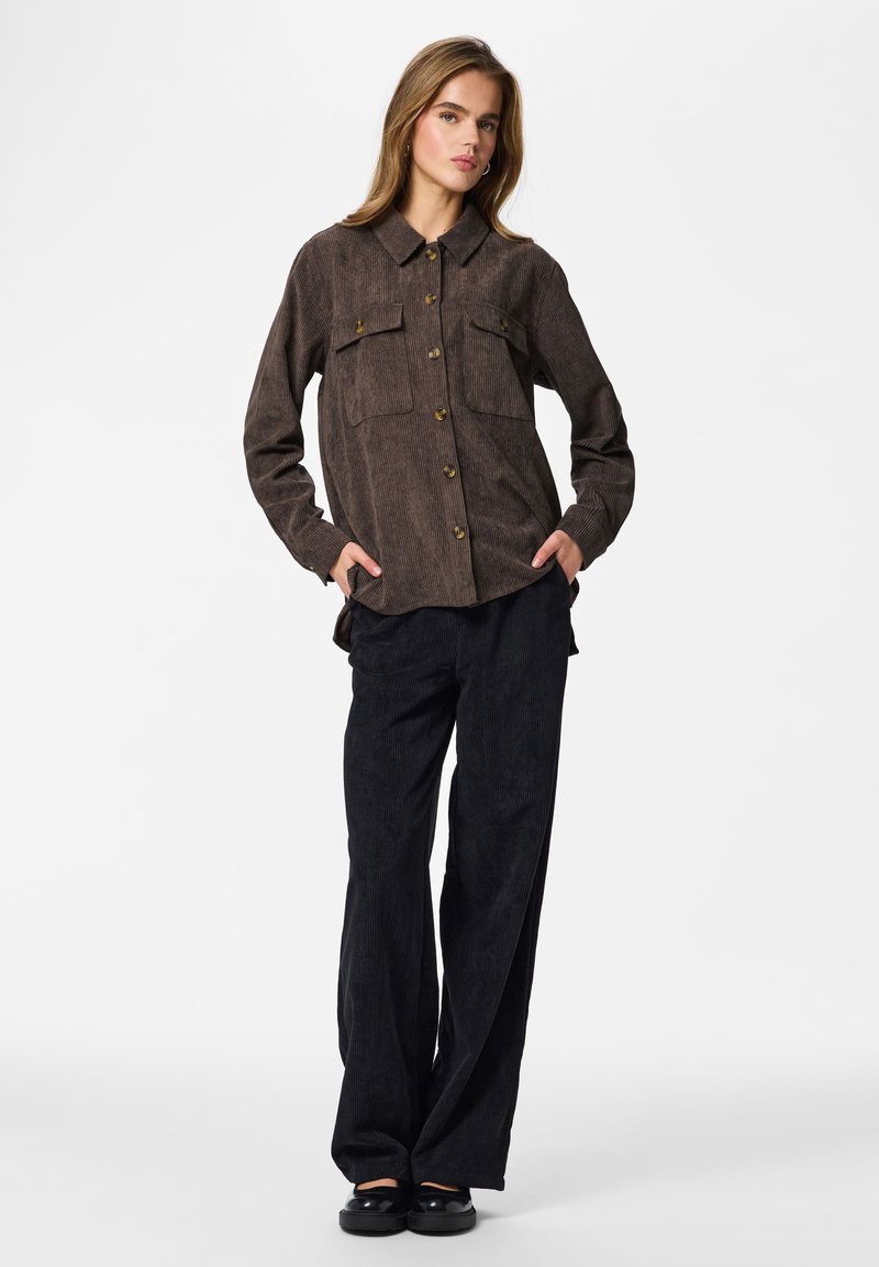 Femme debout, les mains dans les poches, portant une chemise en velours côtelé marron boutonnée, un pantalon noir à jambes larges et des chaussures noires sur un fond uni.
