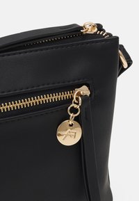 Borsa a mano in pelle nera con zip dorata, hardware e charm con logo. Texture liscia con design elegante e forma minimalista.