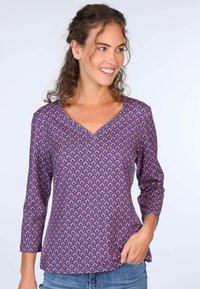 Lila gemusterte Bluse mit V-Ausschnitt und Dreiviertelärmeln. Das Design weist ein sich wiederholendes Muster in roten und blauen Tönen auf. Glatte Stofftextur.