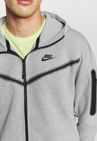 Felpa grigia con zip, dettagli neri e logo Nike sul petto. Presenta un cappuccio, tasche con zip e una trama liscia sul tessuto.