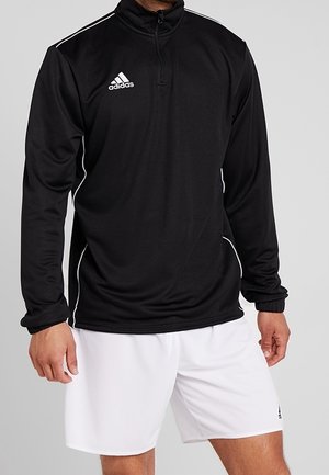 Mies pukeutunut mustaan Adidas pitkähihainen vetoketjulliseen puseroon, jossa valkoiset reunat, ja valkoiset Adidas-shortsit yksiväristä taustaa vasten.