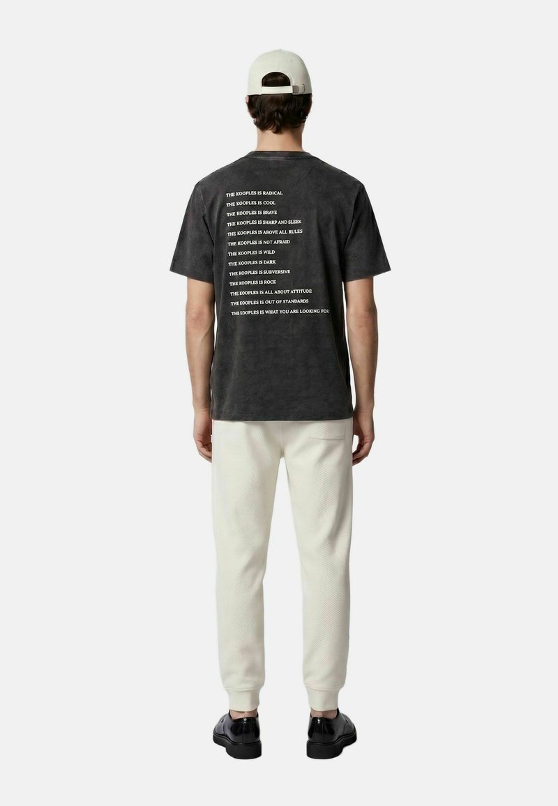 Hombre con gorra blanca, camiseta negra con gráficos y varias líneas de texto blanco en la espalda, pantalones blancos y zapatos negros, de espaldas sobre fondo liso.