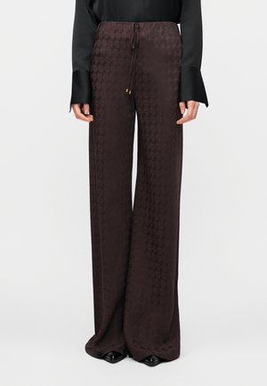 Femme portant un pantalon marron foncé taille haute à jambes larges avec un motif subtilement embossé et une taille à cordon, assorti à des chaussures noires à bouts pointus.