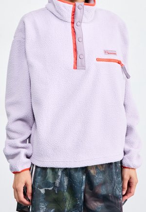 Lawendowy polarowy sweter z koralową plisą zapinana na zatrzaski i kieszenią na piersi zapinaną na zamek, noszony z szortami w kamuflażowy wzór.