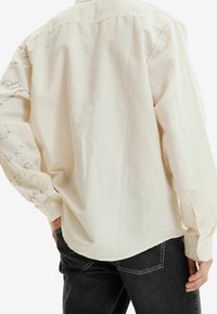 Camicia di colore crema, a maniche lunghe, con una texture morbida e liscia. Presenta un motivo stampato sulla manica sinistra e bottoni sui polsini.