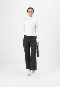 Weißes langärmeliges Rollkragenoberteil, schwarze weitgeschnittene Jeans und weiße Sneakers. Die Mode trägt eine kleine schwarze Handtasche und steht vor einem schlichten Hintergrund.