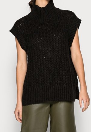 Femme portant un pull col roulé en grosse maille noire sans manches et un pantalon large kaki sur un fond uni.