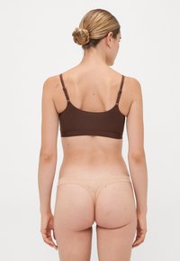 Vrouw met lichte huid en blond haar in een knot, gekleed in een bruine bh en beige strings, van achteren gezien tegen een effen witte achtergrond.
