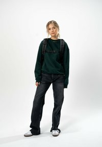 Felpa verde scuro a vestibilità morbida, abbinata a jeans neri a gamba dritta e sneakers bianche. Zaino nero indossato su entrambe le spalle.