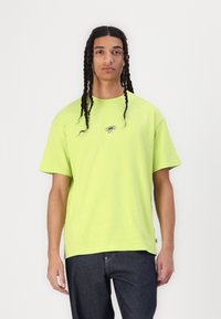 Bright lime green oversized t-shirt made of cotton, featuring short sleeves and a small black logo on the chest. Paired with dark denim jeans.