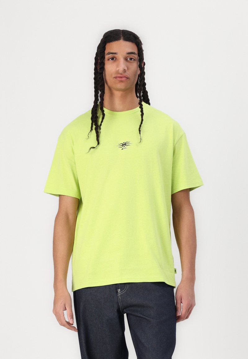 Bright lime green oversized t-shirt made of cotton, featuring short sleeves and a small black logo on the chest. Paired with dark denim jeans.