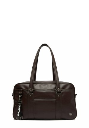 Tmavě hnědá kožená kabelka se dvěma dlouhými uchy, přední kapsou a černým klíčenkou s bílým nápisem připevněnou na levé straně.