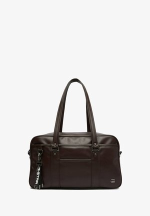 Ciemnobrązowa skórzana torba na ramię z dwiema długimi rączkami, przednią kieszenią oraz czarnym brelokiem z białym napisem przymocowanym po lewej stronie.