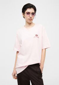 Άτομο που φοράει ανοιχτό ροζ μπλουζάκι Adidas, σκούρο παντελόνι και γυαλιά με μοβ απόχρωση, που στέκεται μπροστά σε ένα απλό λευκό φόντο.